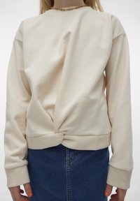 Sweatshirt boje slonovače s čvorastim detaljem na leđima, dugim rukavima i rebrastim manžetama, uparen s plavim traper suknjom.
