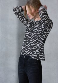 Schwarz-weißer, gestreifter Langarm-Oberteil aus leichtem Stoff, kombiniert mit dunklen Jeans, vor einem grauen Hintergrund.