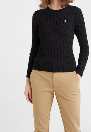 Kobieta w czarnym swetrze w warkocze z małym białym logo i beżowych spodniach, z jedną ręką w kieszeni, na jednolitym tle.