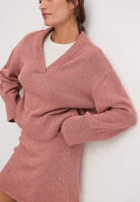 Texturirana roza pletena jopica z globokim V-izrezom in rebrasto manšeto, v kombinaciji z ujemajočo se prilegajočo krilom. Mehka tkanina z vsakdanjim silhueto.