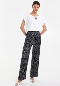 Biała bluzka z wiązanym dekoltem, w połączeniu z czarnymi spodniami z szerokimi nogawkami z wysokim stanem w białe kropki. Modelka ma na sobie białe sandały na obcasie.