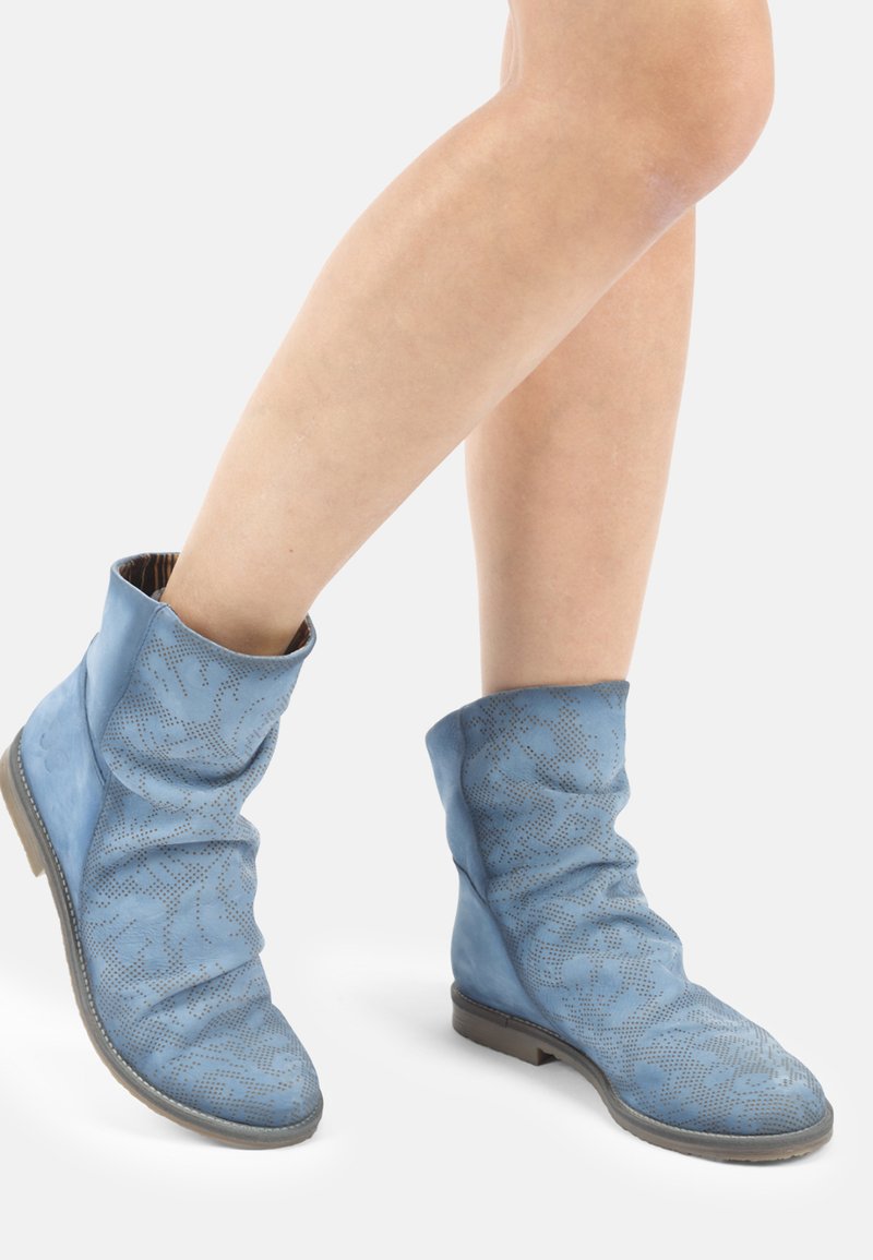 Bottines bleues décontractées à la cheville avec des motifs floraux perforés portées sur des jambes nues sur fond blanc.