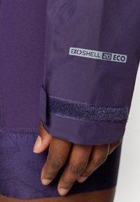 Fialová voděodolná bunda, rukáv se suchým zipem; obsahuje štítek "EXOSHELL 20 ECO". Materiál se zdá být hladký, s texturovaným vzorem pod ním.