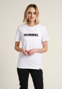 T-shirt en coton blanc avec un logo "HUMMEL" noir audacieux, manches courtes, col rond et une étiquette rouge accentuée à l'ourlet.