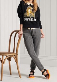 Svart sweatshirt med tiger grafik och "SUPERDRY" text, grå joggers och gula-orange sandaler, med en minimalistisk bakgrund.