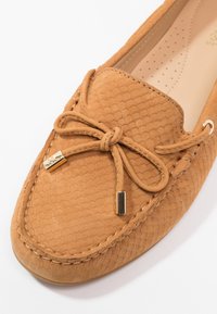 Chaussure mocassin couleur beige clair avec un motif en nid d'abeille texturé, dotée d'un nœud papillon et d'accents métalliques dorés. Matière en daim doux avec des coutures apparentes.