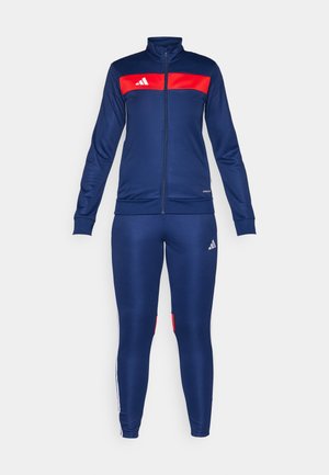 Marineblauwe trainingspak met een rood paneel op de jas, voorzien van een rits en witte strepen op de broek. Gemaakt van soepele, rekbare stof.