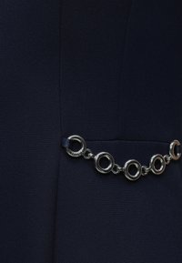 Silbernes rundes Kettendetail, befestigt an dunkelmarine Stoff, das den Design- und Texturkontrast bei der Kleidung hervorhebt.