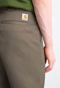 Nærbillede af en person iført brune Carhartt-bukser og en grøn skjorte, der viser baglommen og en synlig underarm med vener.