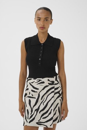 Femme portant un haut sans manches en maille côtelée noire avec des boutons et une jupe portefeuille beige à rayures zèbre noires, debout devant un fond uni.