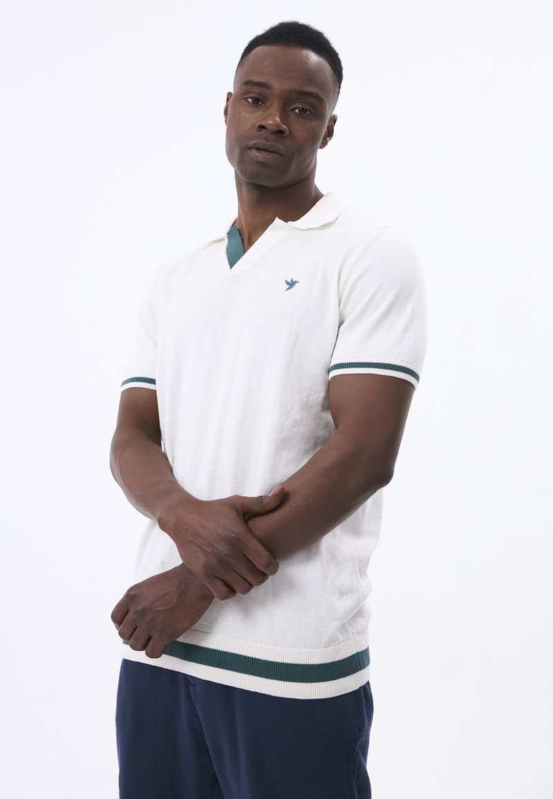 Hvid strikket polo-shirt med en grøn accent på kraven og ærmerne. Har et logo på brystet og et struktureret stof.