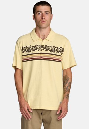 Koszulka polo z lekkiej żółtej bawełny z brązowym kwiatowym wzorem i poziomymi paskami na wysokości klatki piersiowej; posiada krótkie rękawy i klasyczny kołnierz.