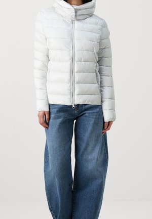 Personne portant une veste matelassée blanche avec un col montant et une fermeture éclair à l'avant, associée à un jean en denim bleu ample, sur un fond uni.