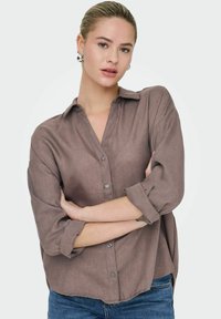 Brun linned skjorte med knapløs krave, lange ærmer der er oprullet, og en afslappet pasform. Har seks knapper og et afslappet design.