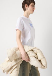Tricou alb cu mâneci scurte, având un logo CK albastru. Modelul ține o jachetă crem cu model matlasat. Pantaloni din denim albastru, cu o croială drept.
