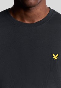 T-shirt nera in cotone con collo a giro e maniche corte. Presenta un piccolo logo di un'aquila ricamato in giallo sul petto. Texture liscia.
