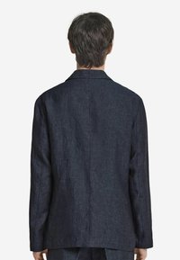 Mörkblå linnakavaj med texturerad tyg, enkel ryggslits, sneddade slag och långa ärmar, sedd från baksidan.