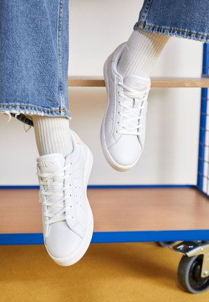Sneakers sintetici bianchi con perforazioni, lacci piatti e marchio Fila. Indossati con calzini leggeri a coste e denim blu, con bordi sfilacciati.