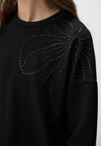 Felpa nera con scollo rotondo e un intricato design di borchie lucide che formano un motivo floreale sul davanti.