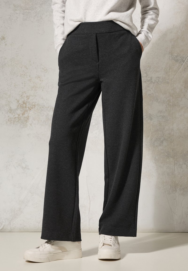 Personne portant un pantalon noir à jambes larges, des baskets blanches et un haut à manches longues gris clair, debout les mains dans les poches contre un mur texturé.