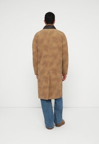 Beige suede coat with a long shape and brown collar. Features a clean back design with a center seam and minimal detailing. Paired with blue jeans.