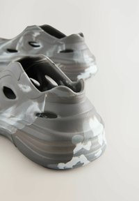 Grå støvler i formet materiale med en tekstureret overflade, der har udskæringer og et hvidt camouflagemønster langs sålen, hvilket fremhæver et unikt design.