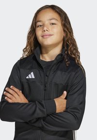 Svart zip-up jacka med hög krage, kontrasterande texturerade paneler och Adidas-logotypen i vitt. Slät tyg med ribbade muddars.