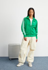 Grön zip-up träningsjacka med vita ränder, kombinerad med lösa krämfärgade byxor och beige sneakers. Texturerad yta och sportig design.