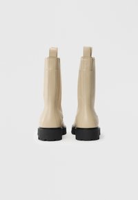 Un paio di stivali in pelle beige con suole spesse nere, mostrati da dietro su uno sfondo bianco.