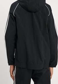 Černá bunda na zip s kapucí, bílé akcentační pruhy podél rukávů a elastické manžety a lem. Lehce, hladký materiál.