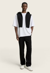 Tricou alb oversized cu pulover negru cu valuri drapat pe umeri, asortat cu pantaloni negri și adidași albi. Design simplu, croială casual.
