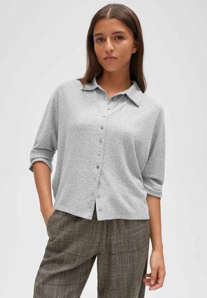 Jonge vrouw met steil bruin haar draagt een lichtgrijs overhemd met knopen en hooggetailleerde bruine geruite broek met handen in de zakken.