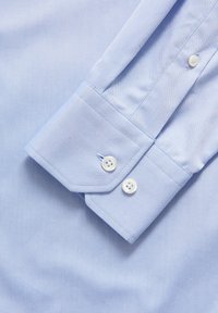 Ljusblå skjorta i ett mjukt tyg, med långa ärmar, spetsiga manschetter och två vita knappar på varje manschett.