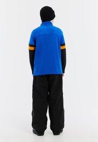 Niebieska koszula z długim rękawem o siateczkowej teksturze, czarne rękawy i pomarańczowy pasek. Luźne czarne spodnie i czarna czapka beanie.