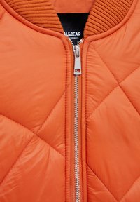 Veste matelassée orange avec une fermeture éclair argentée, un col côtelé et une texture lisse. Logo visible sur l'étiquette intérieure.