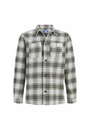 Ung person står med en hvid t-shirt, sort og hvid ternet overshirt, løstsiddende blå jeans og beige sko mod en ensfarvet baggrund.