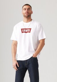 Bela bombažna majica s rdečim logotipom Levi's. Kratke rokave, kroj s krožno ovratnikom, v kombinaciji s temno modrimi kavbojkami za kontrast.