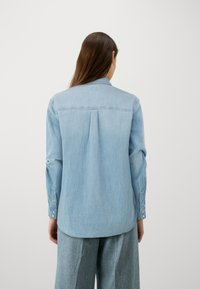 Světle modrá denimová košile s dlouhými rukávy, knoflíkovými manžetami a designem na zadním díle. Tkanina má hladkou strukturu a volný střih.