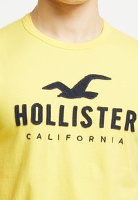Gul bomull t-shirt med en svart broderad logotyp där texten "HOLLISTER CALIFORNIA" finns under en grafik av en mås.