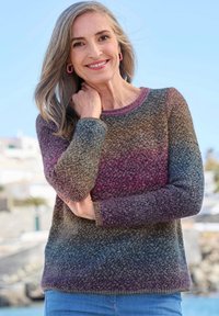 Femme souriante aux cheveux gris portant un pull en tricot dégradé aux nuances de violet, marron et bleu, se tenant à l'extérieur sous un ciel dégagé.