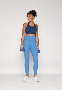 Reggiseno sportivo blu con fascia, abbinato a leggings a vita alta azzurri. Maniglie nere nei pugni e scarpe da ginnastica bianche completano il look.