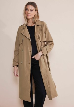 Beige Trenchcoat mit großen Revers, doppelter Knopfleiste und Gürteldetails. Leichte, glatte Stoffqualität. Getragen über einem schwarzen Oberteil und einer Hose.