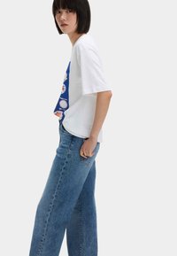 Witte t-shirt met korte mouwen en blauwe grafische print, gecombineerd met lichtblauwe, high-waisted spijkerbroek. Casual pasvorm en eenvoudig ontwerp.