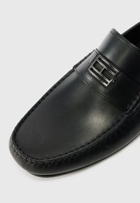 Musta nahkakengät, joissa on sileä pinta, varustettu erottuvalla hopeanvärisellä metallikoristeella ja kontrastivärillä oleva ompelu reunojen ympärillä.