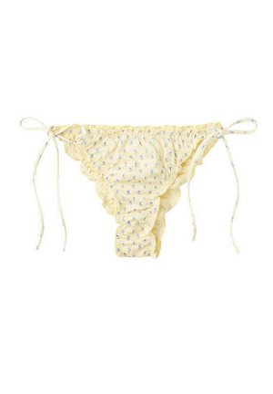 Femme portant un bikini jaune à motifs et des lunettes de soleil noires, debout dans une eau peu profonde et claire avec des collines rocheuses en arrière-plan sous un ciel bleu.