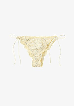 Femme portant un bikini jaune à motifs et des lunettes de soleil noires, debout dans une eau peu profonde et claire avec des collines rocheuses en arrière-plan sous un ciel bleu.