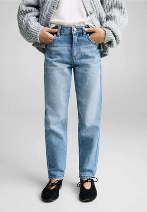 Personne portant un jean taille haute bleu clair, un cardigan en maille gris, un haut blanc et des ballerines noires avec des lacets noués, les mains dans les poches.