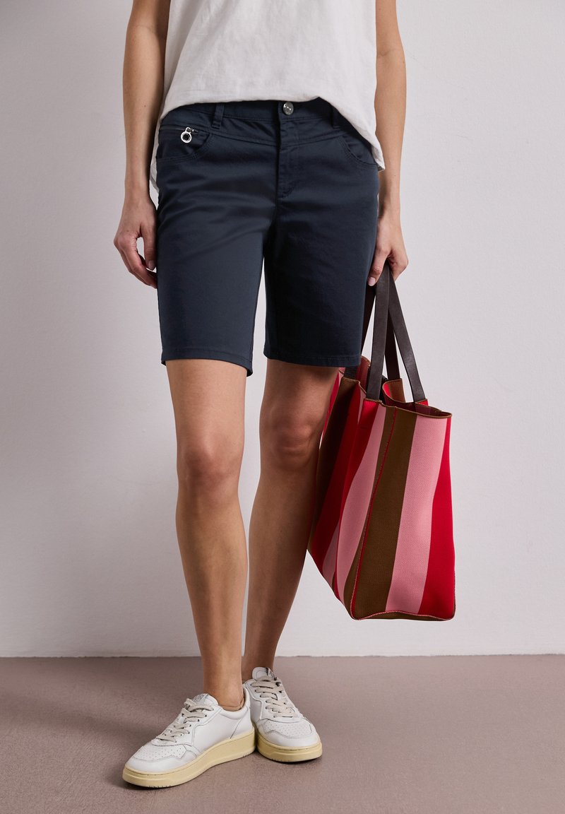 Personne portant un short bleu marine et des baskets blanches, tenant un grand sac cabas rayé rouge, rose et marron avec des poignées foncées.