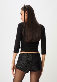 Czarna bluzka z długim rękawem w połączeniu z czarnymi szortami z cekinów o dopasowanym kroju i błyszczącej teksturze, na tle jednostajnego białego tła.