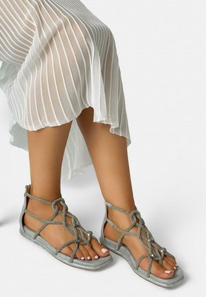 Pieds portant des sandales plates argentées à brides avec des nœuds décoratifs, associées à une jupe blanche plissée et transparente sur un fond uni.